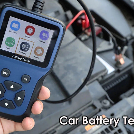 A hand holding a blue and black car battery tester diagnostic tool with a screen showing diagnostic icons, with a blurred car engine bay in the background and the text 'Car Battery Tester' in the bottom right corner.