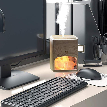 A desktop humidifier with a light wood grain exterior and a glowing orange interior, shown on a desk next to a keyboard, mouse, and computer monitor.
