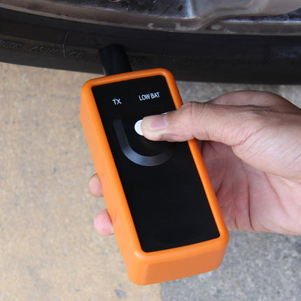Hand holding an orange diagnostic tool with a screen displaying 'LOW BAT' on a car's undercarriage.