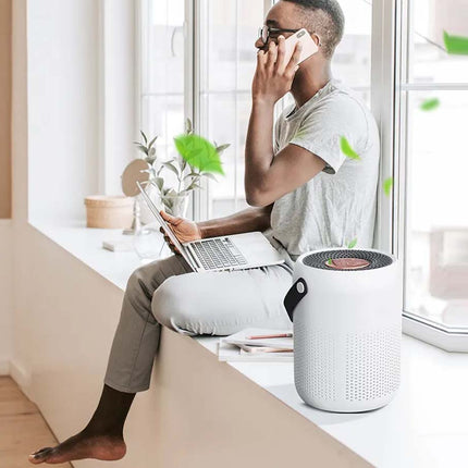 A man sits on a windowsill in a bright room, talking on a phone and using a laptop, with a white cylindrical air purifier placed on the sill beside him.