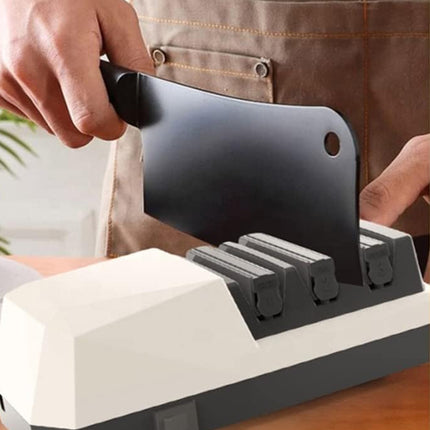 A close-up of a person using a white and dark gray knife sharpening machine, sliding a black cleaver knife into one of the sharpening slots. The person is wearing a brown canvas apron.