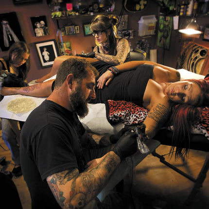 A tattoo artist wearing black gloves and a black t-shirt tattoos a client with red hair on a tattoo table inside a tattoo studio. Another artist is visible in the background working on a client.