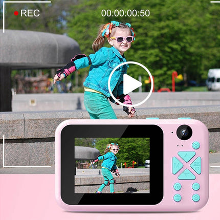 A pink children's digital camera with turquoise buttons is shown in front of a screen displaying a video of a young girl riding a bicycle outdoors. The image includes a camera recording overlay with 'REC' and a video play button.