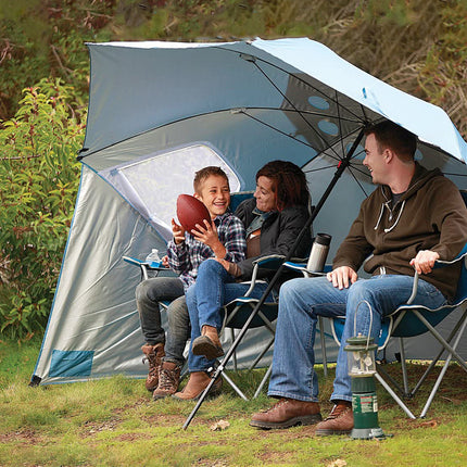 Three people sitting under a large umbrella in a camping setting with trees in the background.