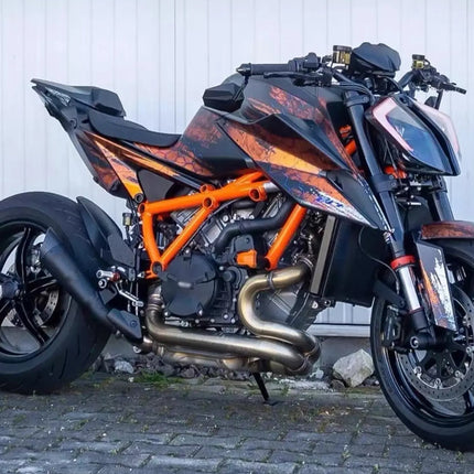 A black and orange KTM motorcycle with a custom flame graphic, parked on a cobblestone surface against a white corrugated metal wall.