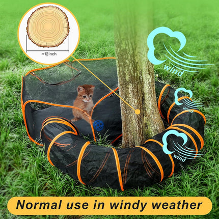 A black and orange cat play tunnel set up on grass next to a tree, with a small kitten inside. The image includes a circular inset showing the tree-slice size detail, blue wind icons with the word 'wind,' and a yellow text banner reading 'Normal use in windy weather.