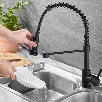 A black pull-down kitchen faucet with a flexible gooseneck spout is shown installed in a stainless steel sink, with a person's hands running water while washing a white plate.