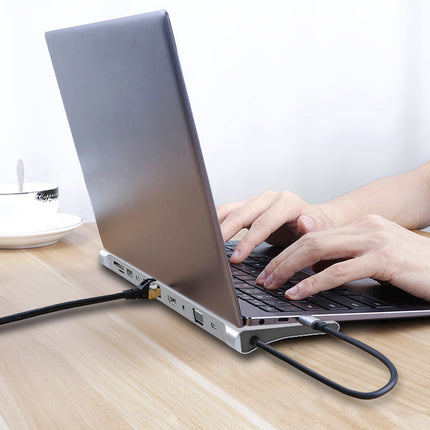 A silver laptop on a light wood desk with a person's hands typing on the keyboard and a black cable plugged into the side port. A white cup and saucer are visible in the background.