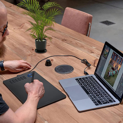 Person using a stylus on a graphics tablet with a laptop displaying a video call on a wooden table.