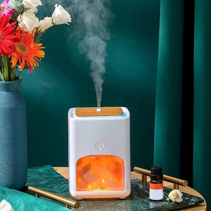 A white and wood diffuser with a glowing orange interior and misting top, styled on a table with a blue vase of flowers, a small essential oil bottle, and dark green curtains in the background.