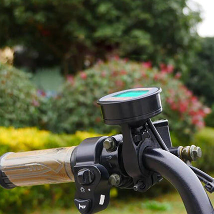 A close-up of a black motorcycle handlebar-mounted display screen with a green-tinted face, attached to a black handlebar clamp. The handlebar grip is tan, and the background is a blurred green garden with pink flowers.