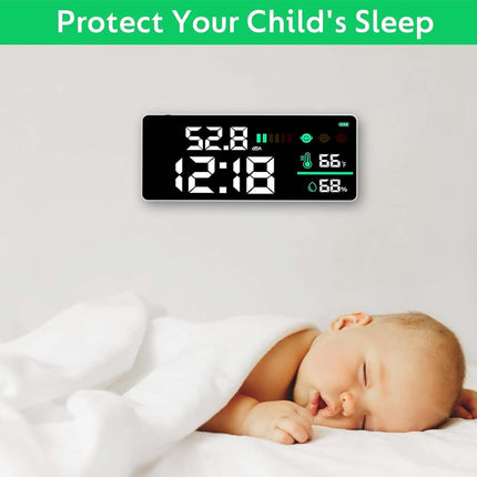 A digital baby monitor and thermometer displayed above a sleeping baby wrapped in a white blanket. The monitor shows the time as 12:18 and body temperature and humidity readings.