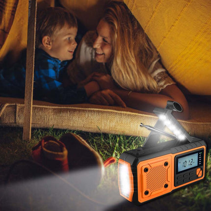 An orange and black portable emergency radio with built-in speakers and a built-in LED flashlight, shown in a camping scene with a child and woman hiding under a tent while the radio illuminates the grass.