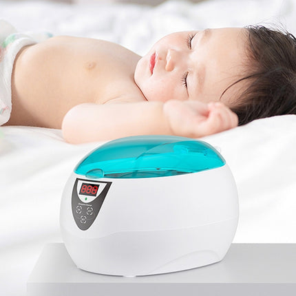 A baby sleeping on a white bed next to a white and turquoise electronic baby bath or foot bath device with a digital display screen.