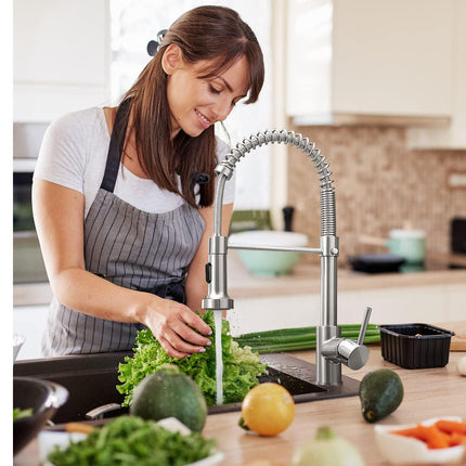 A woman in a kitchen wearing a gray and white striped apron, using a modern pull-down faucet to rinse fresh green vegetables at the sink.