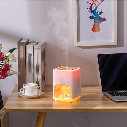 A pink and white cube-shaped humidifier with a glowing orange interior sits on a wooden desk, emitting a visible plume of steam. The scene includes a laptop, books, a cup and saucer, a vase of flowers, and framed wall art in a home office setting.