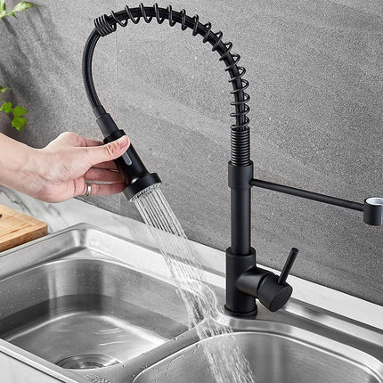 A black gooseneck pull-down kitchen faucet with a flexible spring arm, shown with water running from the spray head into a double-basin stainless steel sink. A hand is holding the spray head.