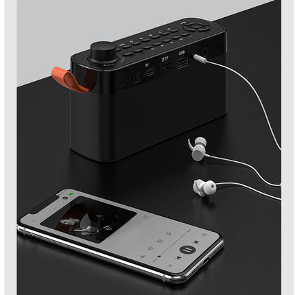 A black portable Bluetooth speaker with a rotary dial and orange accent, shown with a smartphone and white earbuds on a dark tabletop.