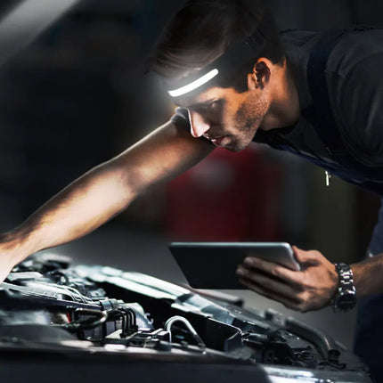 Mechanic using a tablet under the hood of a car with a headlamp illuminating the area.