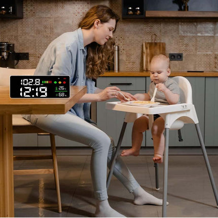 A woman feeding a baby seated in a white high chair in a kitchen, with a digital timer device shown in the foreground displaying time and battery status.