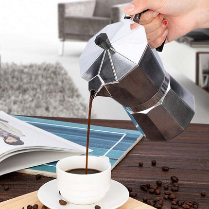 Stovetop espresso maker pouring coffee into a white cup on a wooden surface with coffee beans scattered around.