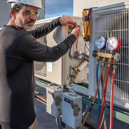 A HVAC technician wearing a white hard hat and safety glasses works on an outdoor air conditioner unit on a rooftop, using a multimeter and connected refrigerant gauges and hoses.