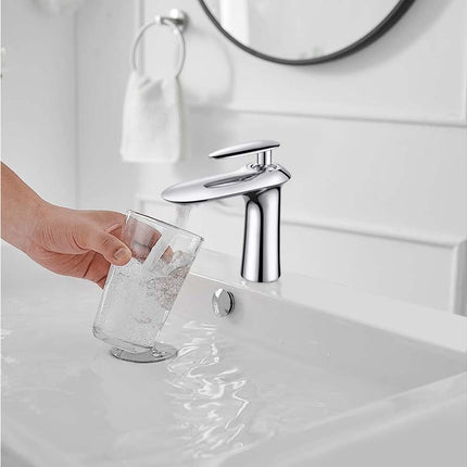 A modern chrome bathroom faucet running with water into a white sink, while a hand holds a clear glass cup under the stream.