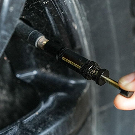 A close-up of a tire valve stem with a person's fingers holding a small black and gold valve tool against the tire.