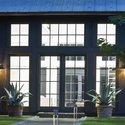 Modern dark charcoal exterior wall with large multi-pane windows, double glass doors, two lit wall-mounted lanterns, potted agaves, and a blue metal roof.