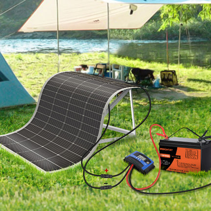 A portable solar panel kit with a flexible black solar panel, a black and orange battery, and a blue digital charger, displayed on a green grassy lawn with a lake, tent, and canopy in the background.