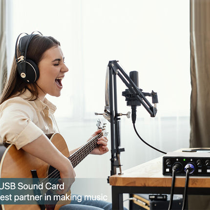 Person playing guitar and singing into a microphone with a USB sound card in a home studio setting.
