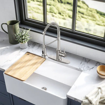 A modern kitchen scene featuring a high-arc chrome pull-down faucet installed in a white farmhouse sink with a white marble countertop, blue-gray cabinets, and a large window showing a green outdoor view.
