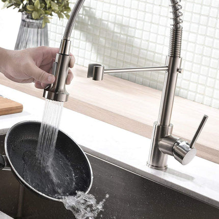 A modern pull-down kitchen faucet with a high-arc spout and side arm, shown with water running from the sink while a hand holds a black frying pan under the stream. The faucet has a brushed stainless steel finish and is installed in a white sink with a light wood countertop and tiled backsplash.