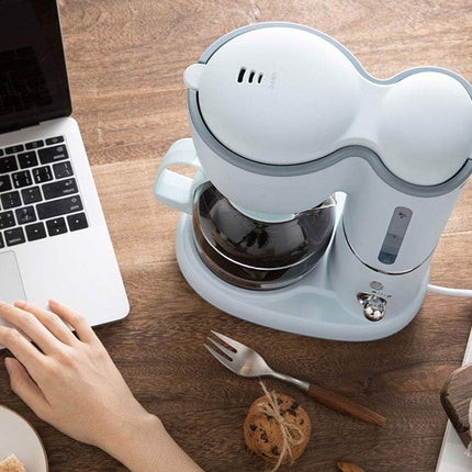 A light blue and gray electric coffee maker sits on a wooden table next to a laptop, with a person's hand resting on the keyboard. A glass carafe filled with coffee is visible, along with a fork, cookies, and a small plate in a styled workspace setting.