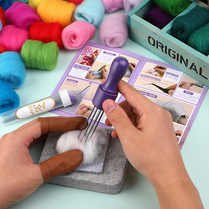 A person's hands are shown using a purple-handled fiber tool on white wool fibers over a gray mat, with colorful balls of yarn, an instruction booklet, and a box of yarn in the background.