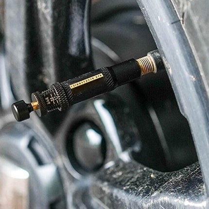 A close-up of a black and gold tire pressure gauge inserted into the valve stem of a black vehicle tire.