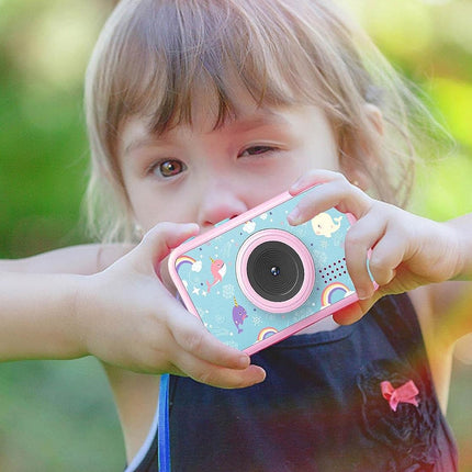 A young child holding a small pink and light blue toy camera with a colorful pattern, photographed outdoors against a soft green bokeh background.