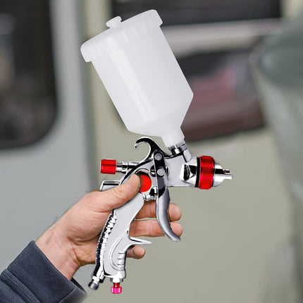 A hand holding a silver and white spray paint gun with red accents and a translucent white paint cup, set against a blurred gray background.