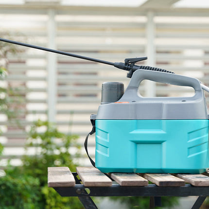 A portable electric pressure washer with a turquoise and gray body sits on a wooden table outdoors, with its long black spray wand extended to the left. A small potted succulent is on the right, and a blurred patio background is visible.