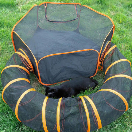 A black and orange cat playpen set on green grass, featuring a tunnel and a mesh play area with a black cat lying inside.