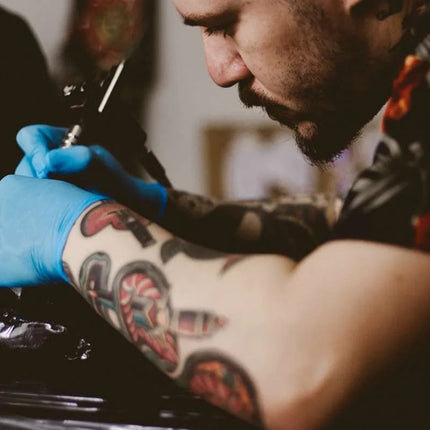 A tattoo artist wearing blue gloves applies a tattoo to a client's arm using a tattoo machine in a dimly lit studio setting.