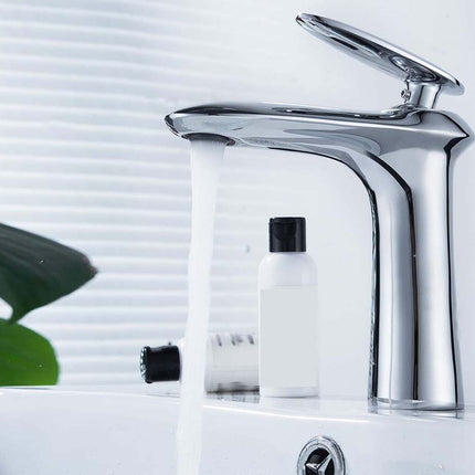 A modern chrome single-handle bathroom faucet with running water, shown installed on a white sink with toiletry bottles and window blinds in the background.
