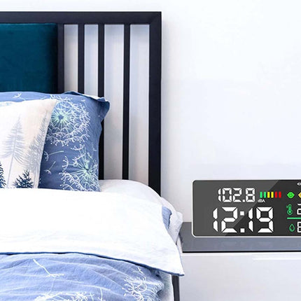 A digital nightstand clock with a large LED display showing the time, battery levels, temperature, and humidity, styled on a dark floating side table next to a bed with blue and white bedding.