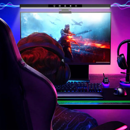 A person seated in a gaming chair facing a computer monitor in a neon-lit home office setup. The desk includes a keyboard, mouse, headset on a stand, and a slim device, with purple, pink, and blue ambient lighting and a glowing RGB desk mat.