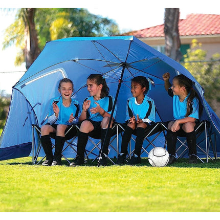 Children sitting under a large blue umbrella in a grassy area with a soccer ball on the ground.
