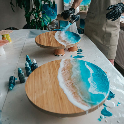 Person applying blue resin to a wooden board on a table with art supplies.
