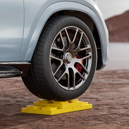 A close-up of a car wheel resting on a bright yellow block-style tire lift or jack placed on a rocky desert surface, with a blurred mountainous background.