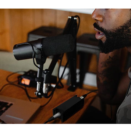 Man speaking into a microphone with headphones on, in front of a laptop on a wooden desk.