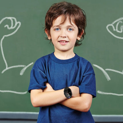 A young boy with brown hair and blue eyes stands with arms crossed in front of a green chalkboard. He is wearing a dark blue crewneck t-shirt and a black smartwatch.