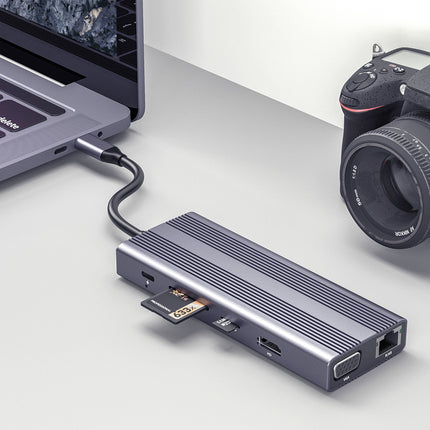 A portable HDMI laptop adapter in a dark metallic gray finish, shown connected to a silver laptop on the left and a black DSLR camera on the right, set against a clean light gray background.